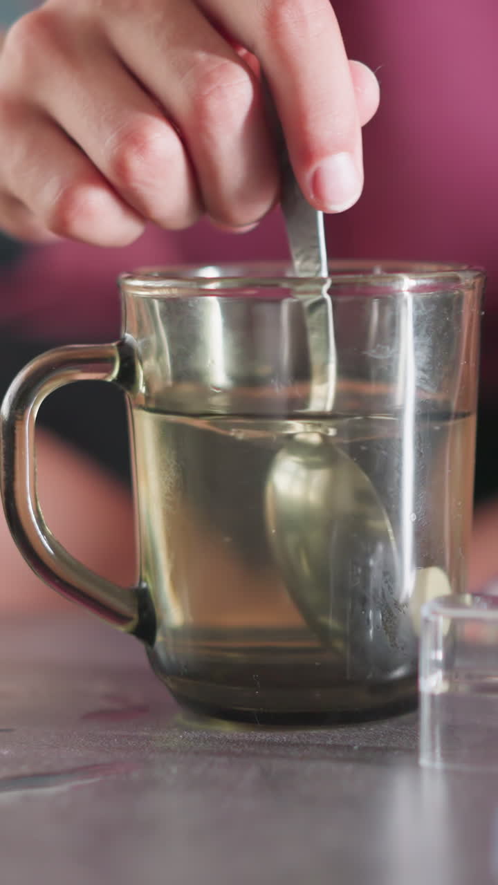 Close up of hand stirring drug in glass of warm water using metal spoon on table with medicine packs and thermometer nearby in calm indoor setting