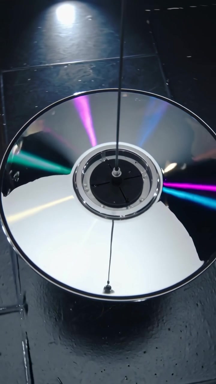 Pystyvideo: Pyörivä CD-levy saa vauhtia, luoden värikaaria mustien laattojen ylle, johdon solmu heiluu.