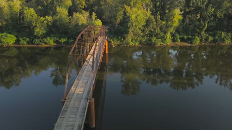 Right-to-left-aerial-slow-truck-shot-of-an-old-iron-bridge-over-a-tree-lined-rural-river-in-warm-late-afternoon-light