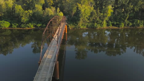 toma aérea de derecha a izquierda de un viejo puente de hierro sobre un río rural bordeado de árboles en la cálida luz de la tarde