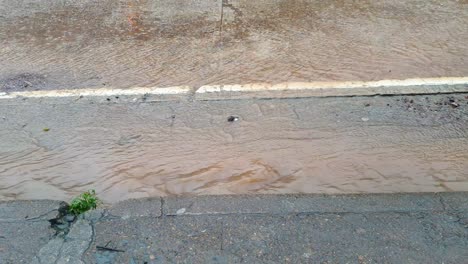 Close-up-of-floodwater-flowing-down-gutters-on-road-with-heavy-raining-downpour-during-monsoon-season