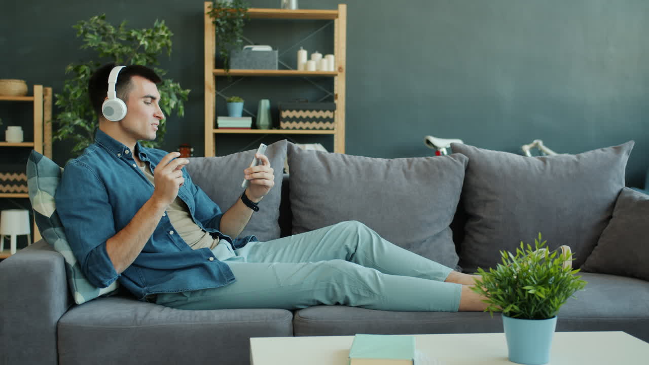 Man Relaxing on a Sofa, Listening to Music and Using a Smartphone