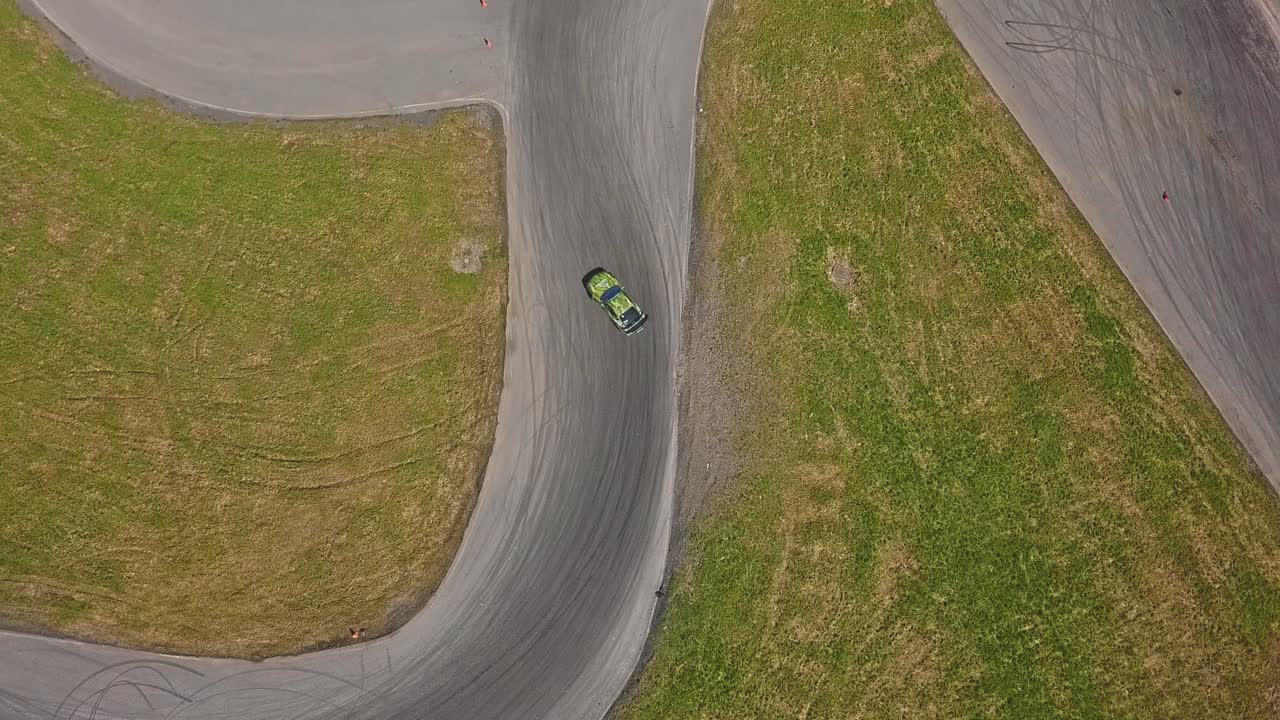 Luftfoto af en bil, der drifter i høj fart i et sving, grønt græs og en racerbane, set oppefra.