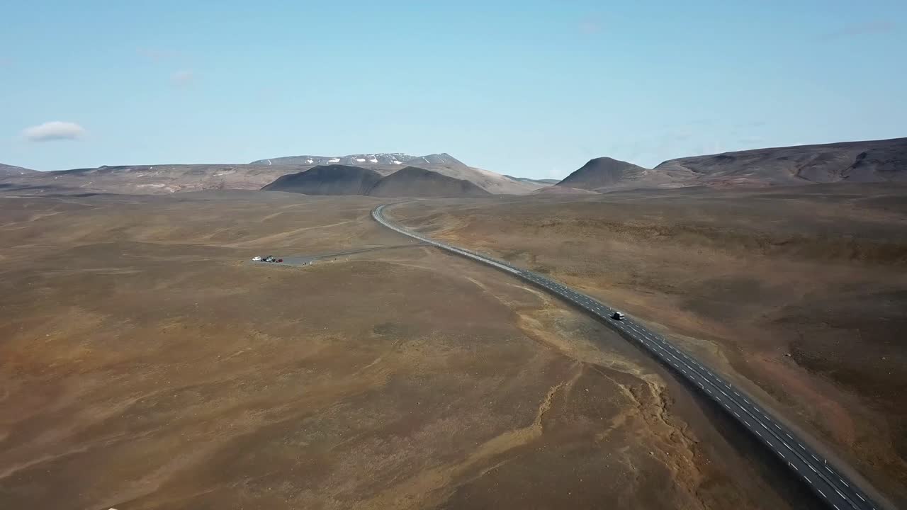 Expansive view of a winding road cutting through the barren, rugged terrain near Parking de la Chaise Blanche, Iceland. Surrounded by distant mountains under a bright blue sky.
