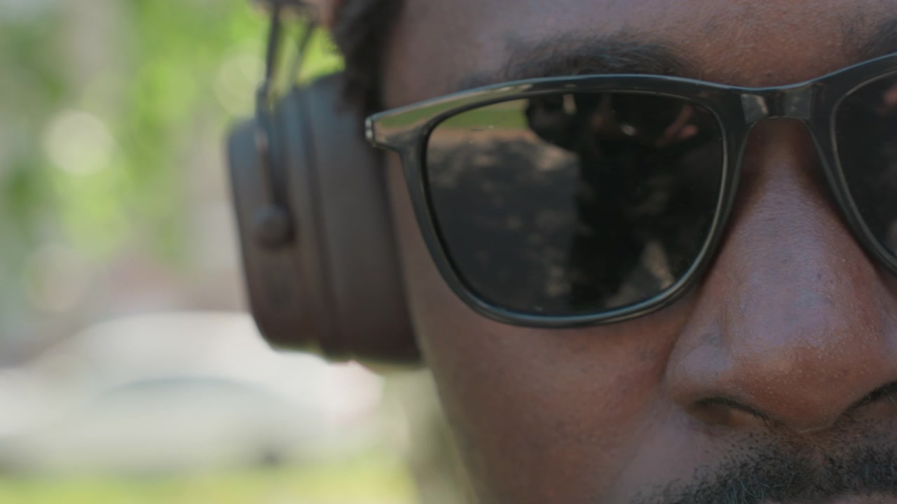 Close up of man wearing sunglasses and ear protection with slight camera movement. reflection in glasses shows another man holding object suggesting interaction or shared activity. Soft