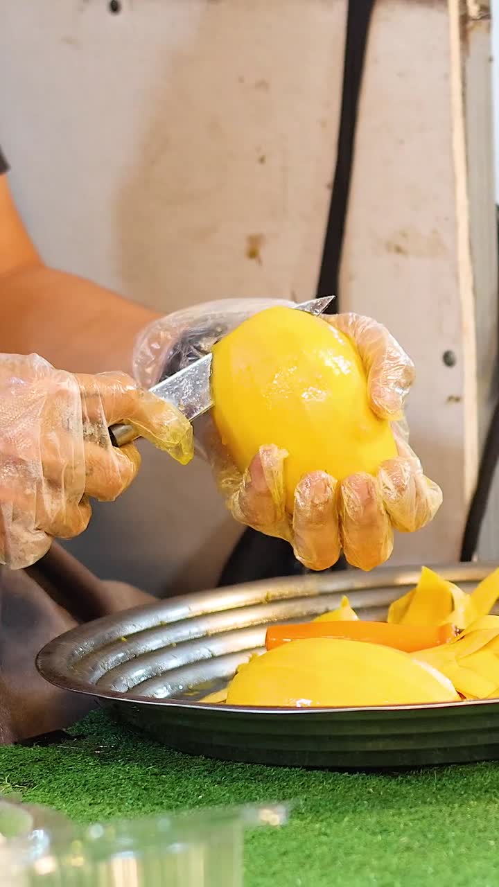 Un venditore di cibo da strada, che indossa guanti di plastica, affetta abilmente un mango giallo maturo a pezzi.