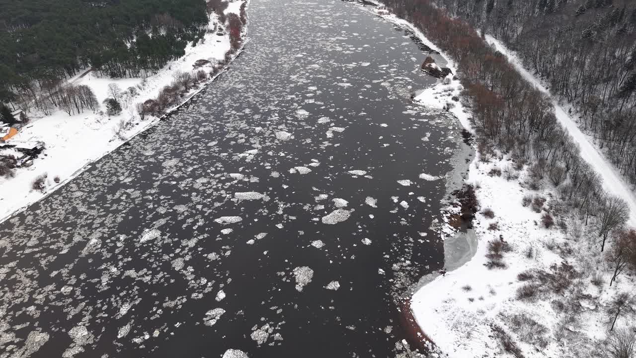 雪に覆われた森林を川が流れる、穏やかな冬の風景を紹介する空撮映像。水路には氷が浮かび、白い背景との印象的なコントラストを生み出しています。