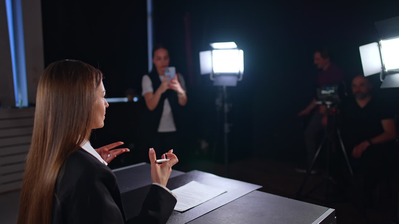 Brunette long-haired lady in black jacket sits in the light of soffits. Cameraman and assisting girl taking video on her phone are at backstage. Blurred backdrop.
