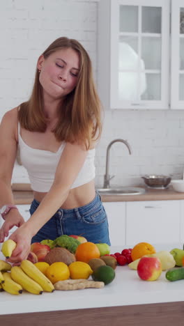 happy young female in the kitchen. beautiful lady throws away centimeter and takes banana. fresh fruit and vegetables on the table. healthy diet. vertical video