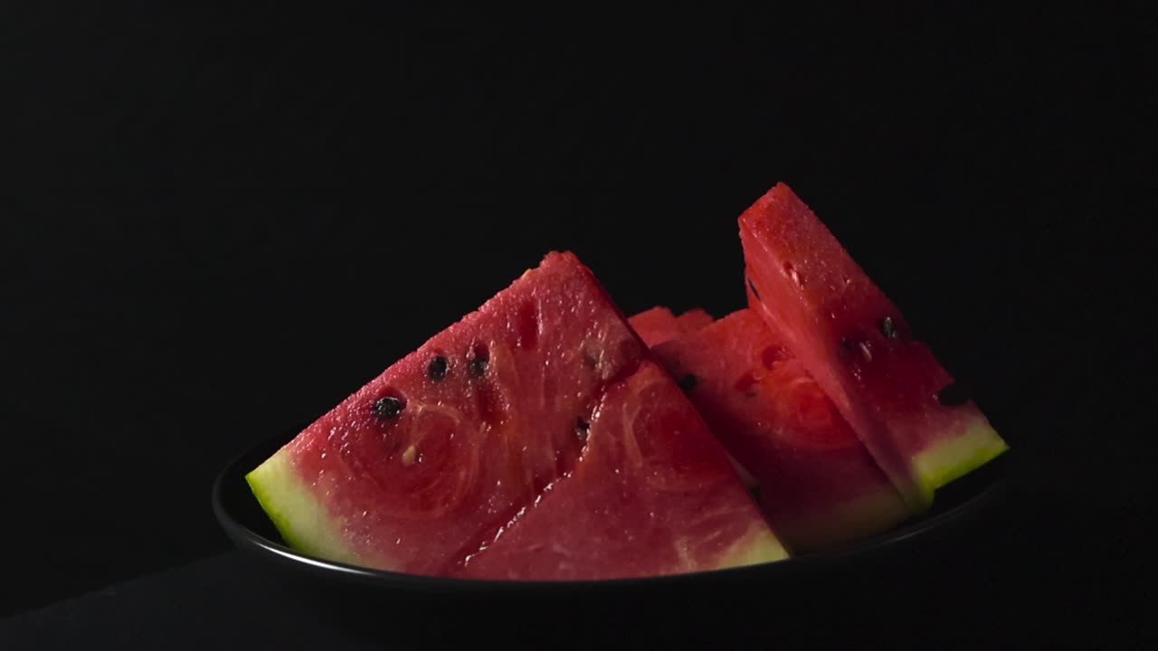 Orbite douce en studio autour de tranches de pastèque triangulaires vibrantes avec des graines visibles dans un bol bleu foncé sur un élégant fond noir