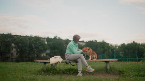 coach seated outdoors on garden bench feeding bulldog dog food while gently petting dog during peaceful morning session in grassy park with clear sky and distant playground structures in background