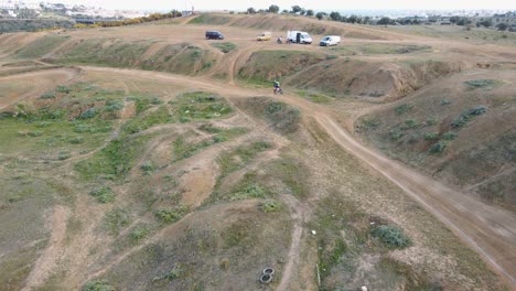 Toma-Aérea-De-La-Pista-De-Motocross-En-Malaga-España-Con-Pista-Polvorienta-Y-Espectadores-En-Las-Colinas-Mientras-Un-Motociclista-Da-Un-Giro-Y-Realiza-Un-Truco-Peligroso