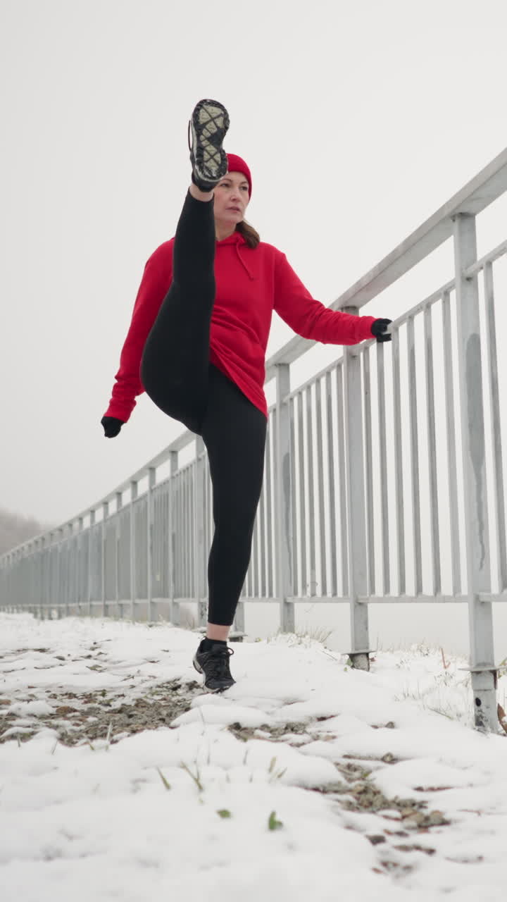 lady exercising outdoors during winter holding iron railing for balance while stretching leg back and forth on snowy ground with foggy atmosphere, light poles and distant river