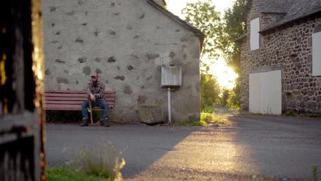 Peaceful-lumberjack-sitting-on-a-bench-in-a-countryside-village-with-old-stone-houses,-at-sunset-with-beautiful-sun-flares