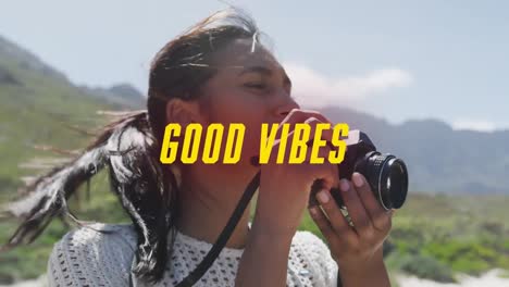 animation of the words good vibes written in yellow over happy woman taking photos in mountains