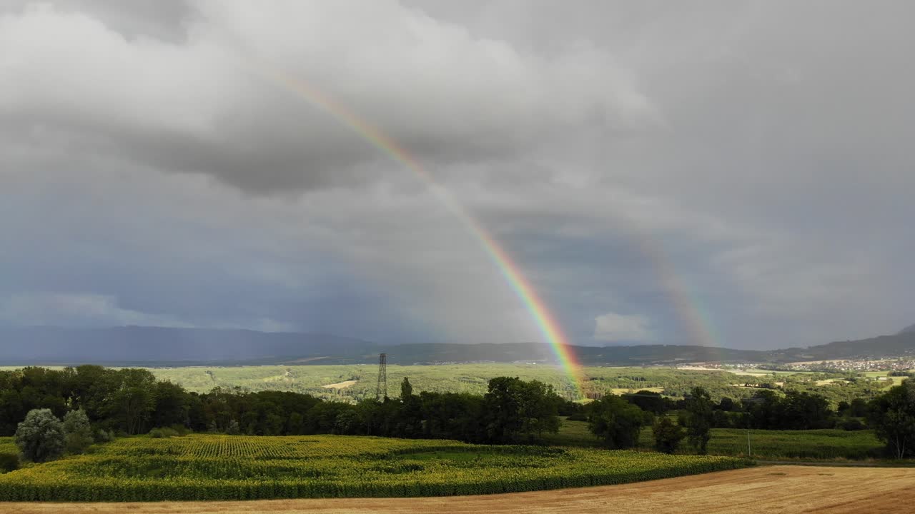 Aerial footage of a rural area in France featuring rolling hills, cultivated fields, a small village, and a vivid rainbow stretching across the sky.
