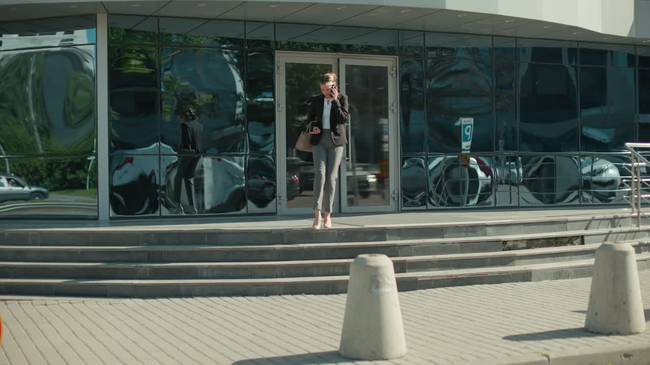 Businesswoman exits modern office building on phone after meeting with client, stepping down staircase while looking to right, holding handbag, with glass facade reflecting cars