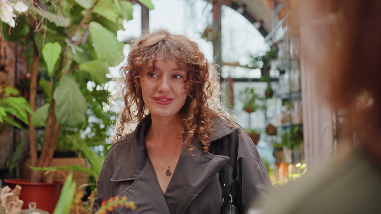 Donna sorridente, cliente in fioreria, ammira un bouquet pastello. Luce solare filtra da una finestra ad arco, piante e vasi sugli scaffali. Conversazione spontanea con la fiorista, risate gentili e atmosfera di vendita.