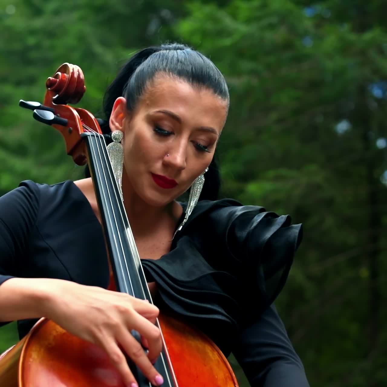 Cello in woman's hands outdoors. Beautiful woman with earrings and black dress playing the musical instrument on nature background.