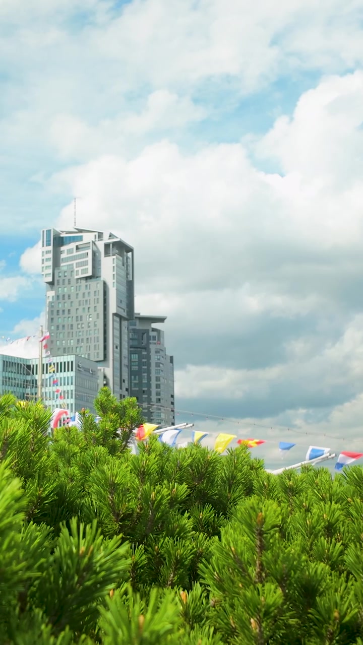 Uma tomada em ângulo baixo das icônicas Torres do Mar em Gdynia, Polônia, emolduradas por exuberantes galhos de pinheiro verde e bandeiras náuticas coloridas sob um céu azul nublado.