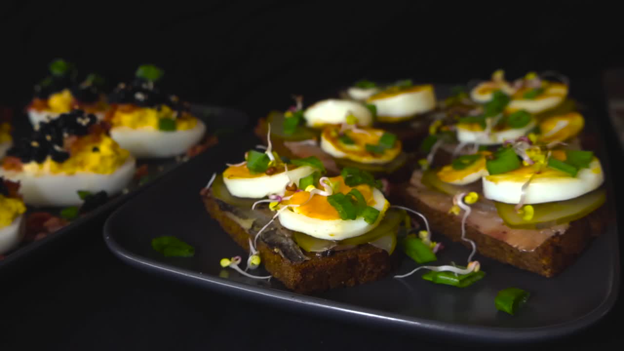 Cinematic slider dolly close view of luxury deviled eggs with black caviar and traditional Baltic sprat sandwiches on rye black bread. Iconic dish served as festive appetizers. Dark blur background