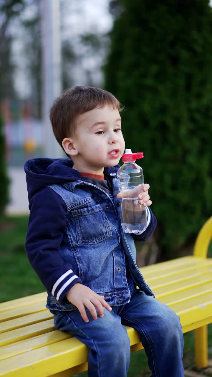 Lovely child sitting on the bench holds the bottle. Kid holds the bottle at his mouth but doesn't drink. Vertical video.