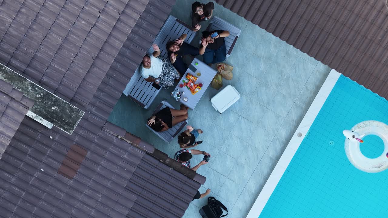 Aerial top-down view of friends waving while enjoying snacks on a patio by a pool