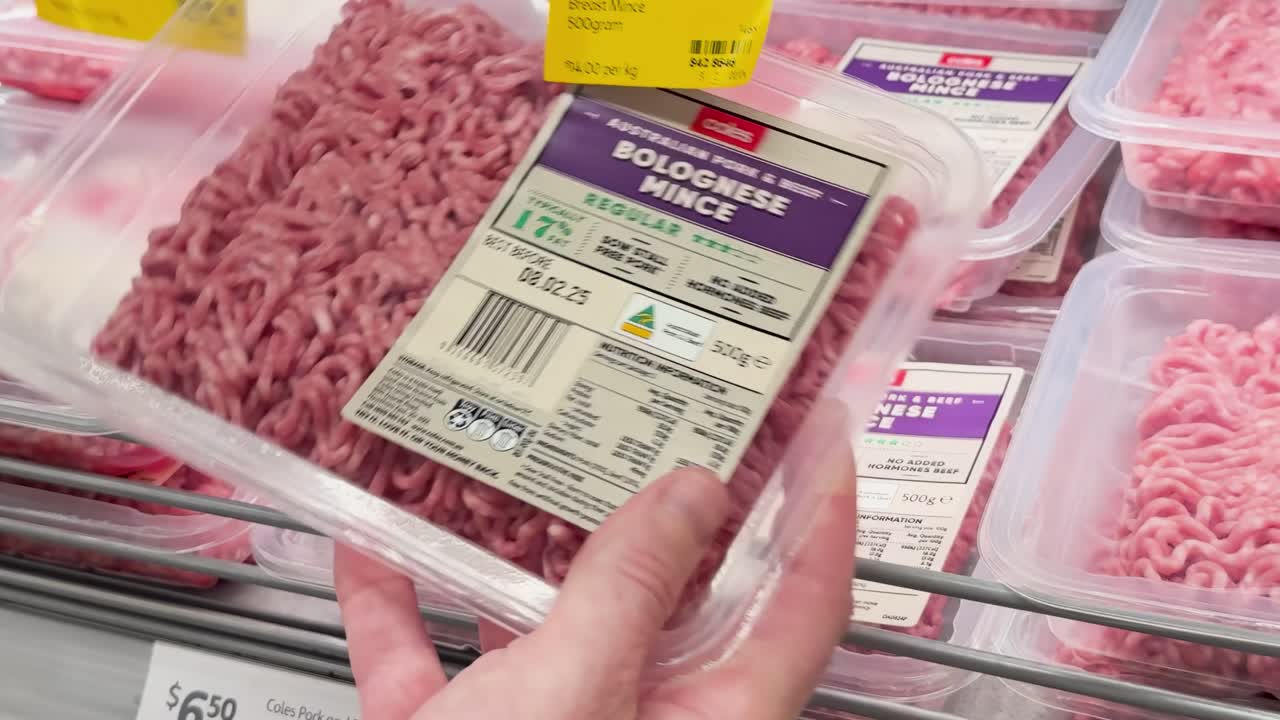 Close-up of hands examining minced meat packages with labels and price tags on a supermarket shelf.