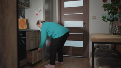 Elderly woman in green sweatshirt bending down to open refrigerator in modern kitchen. A cozy, well-decorated home setting with wooden accents, plants, and soft lighting, highlighting daily routine