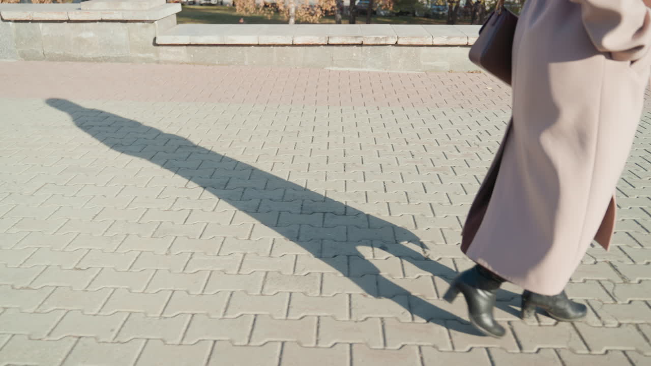 Long coat silhouette casting long shadow across paved plaza, slow stride capturing crisp sunlight, handbag swing, textured tiles, autumn curb, cinematic calm mood, anonymous pedestrian moving