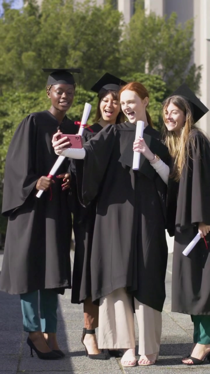 Vertical video: Walking 4 gowned grads pulling pink phone for selfie celebrating diplomas on quad