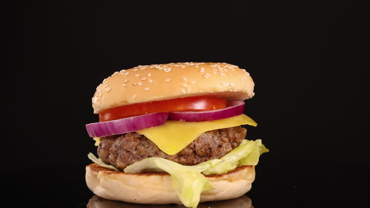 A juicy beef cheeseburger with lettuce, tomato, onion, and cheddar cheese is slowly zoomed in on, under bright studio lighting, with a seamless black backdrop