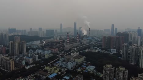 aerial view of industrial complex surrounded by urban landscape, emitting smoke into hazy sky with skyscrapers in background, showing pollution and urban development