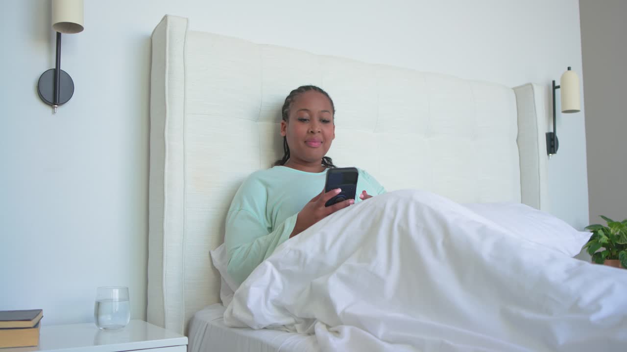 African American woman in bed grasping water glass, checking phone, holding duvet, warming hands
