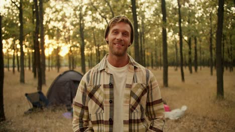 retrato de un tipo con cabello rubio con rastro que se para y mira a la cámara contra el telón de fondo de una tienda y otros participantes en una caminata en un bosque verde soleado de otoño. hombre feliz en una camisa a cuadros posando mientras está de vacaciones en el bosque de otoño
