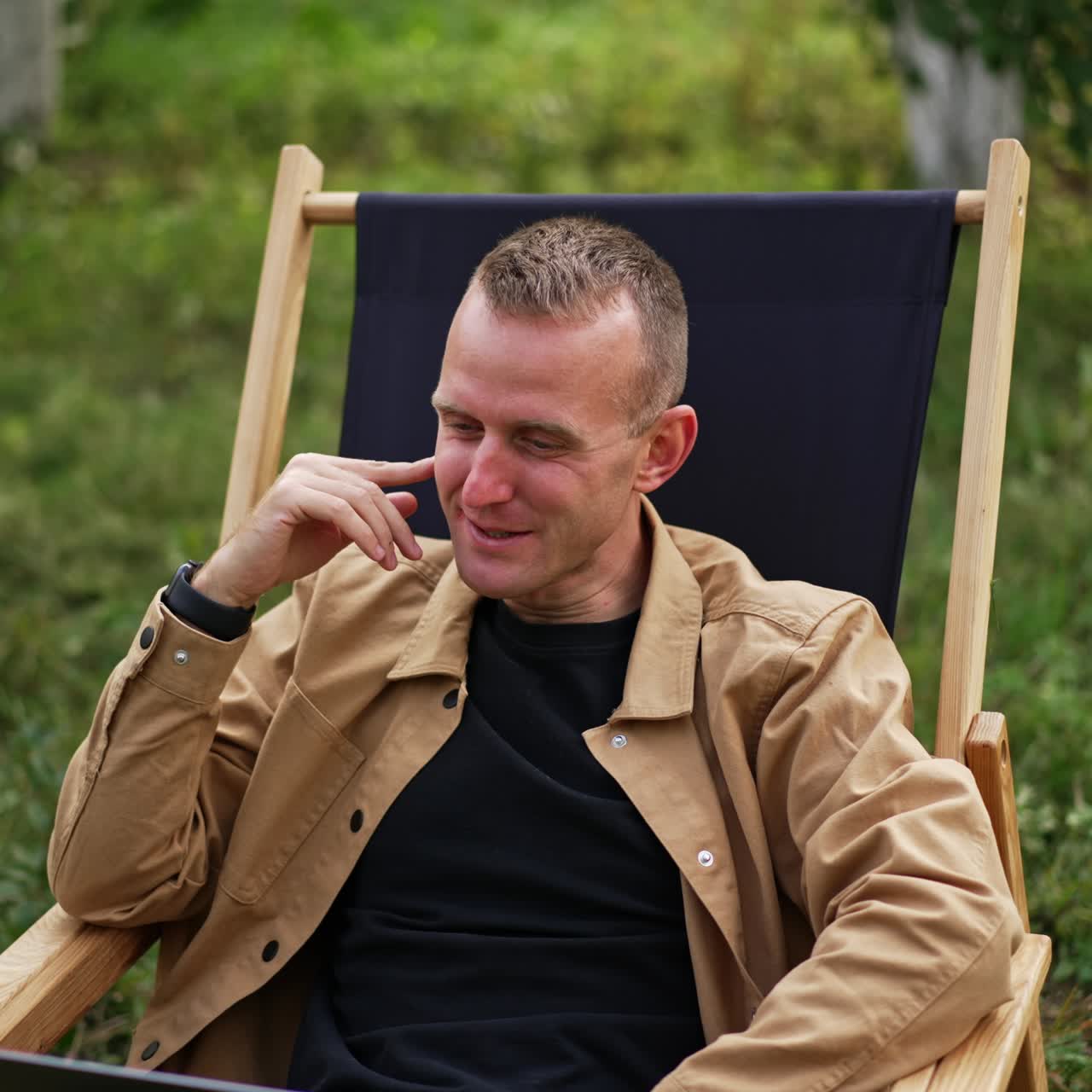 Caucasian man in brown shirt sitting with laptop on his laps in the garden. Relaxed freelance worker talks to someone laughing cheerfully