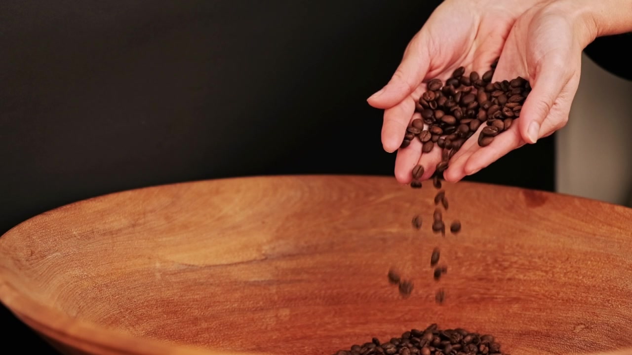 Una donna tiene chicchi di caffè tostati scuri nelle mani e li versa in una grande ciotola di legno.