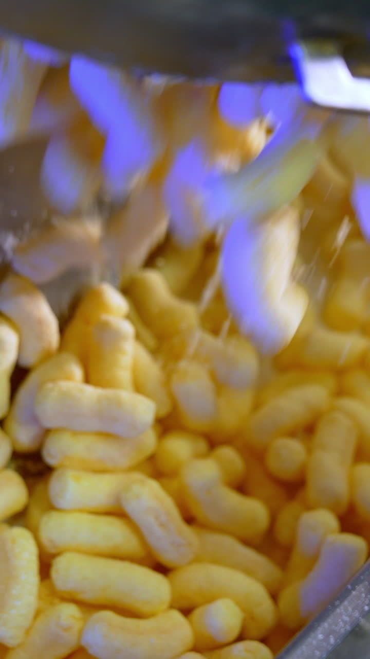 Hand with ladle pushes yellow sweet corn snacks along the conveyor. Close up. Sweet maize sticks production process. Vertical video