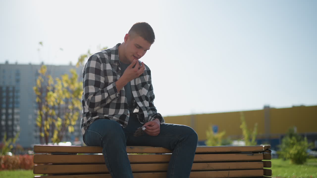young dancer in checkered shirt sits alone on wooden bench outdoors taking medication with blurred view of office buildings parked vehicles greenery and signpost behind under bright morning sunlight