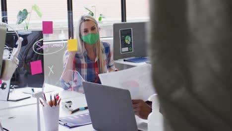 Mixed-race-businessman-wearing-face-mask-sitting-going-through-paperwork-in-modern-office