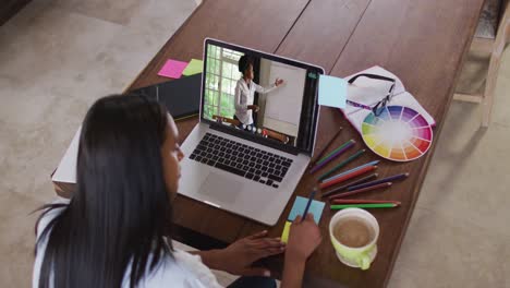 mujer afroamericana escribiendo notas de memoria mientras tiene una videollamada en una computadora portátil en casa