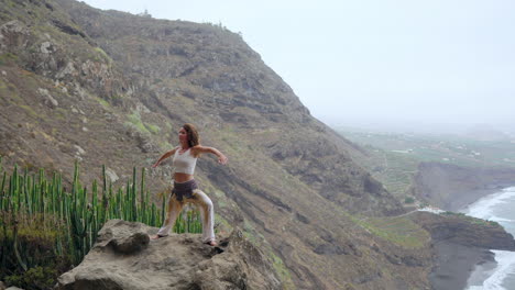 on the shore near rocky mountains, a woman practices yoga's warrior pose, meditating amidst the ocean's expanse—a portrayal of motivation and inspiration in fitness