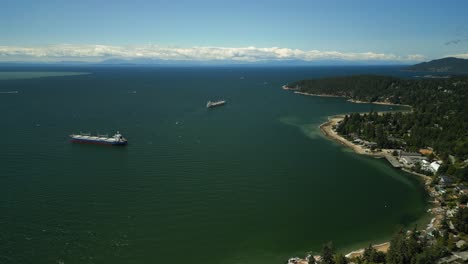 drone flying around the coast line of west vancouver showing the blue ocean water, oil tanker industrial ships anchored in the water, green forests on the hills and bowen island in the background