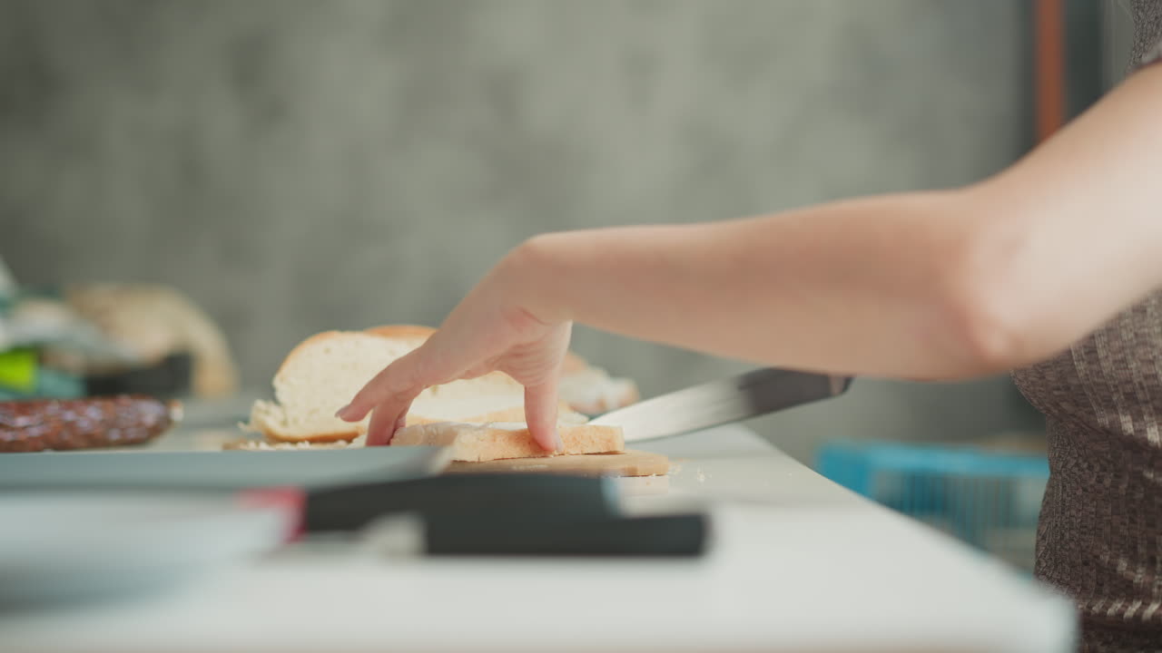 Culinary artist cutting bread into bite sized pieces on wooden board, arranging pieces beside sausage banana eggs and bowl on white table, capturing calm cooking preparation scene