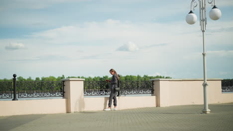 woman in grey clothing leaning against iron railing while reading a book at the riverfront, she carries a black handbag, with trees visible in the background, with a decorative lamp post