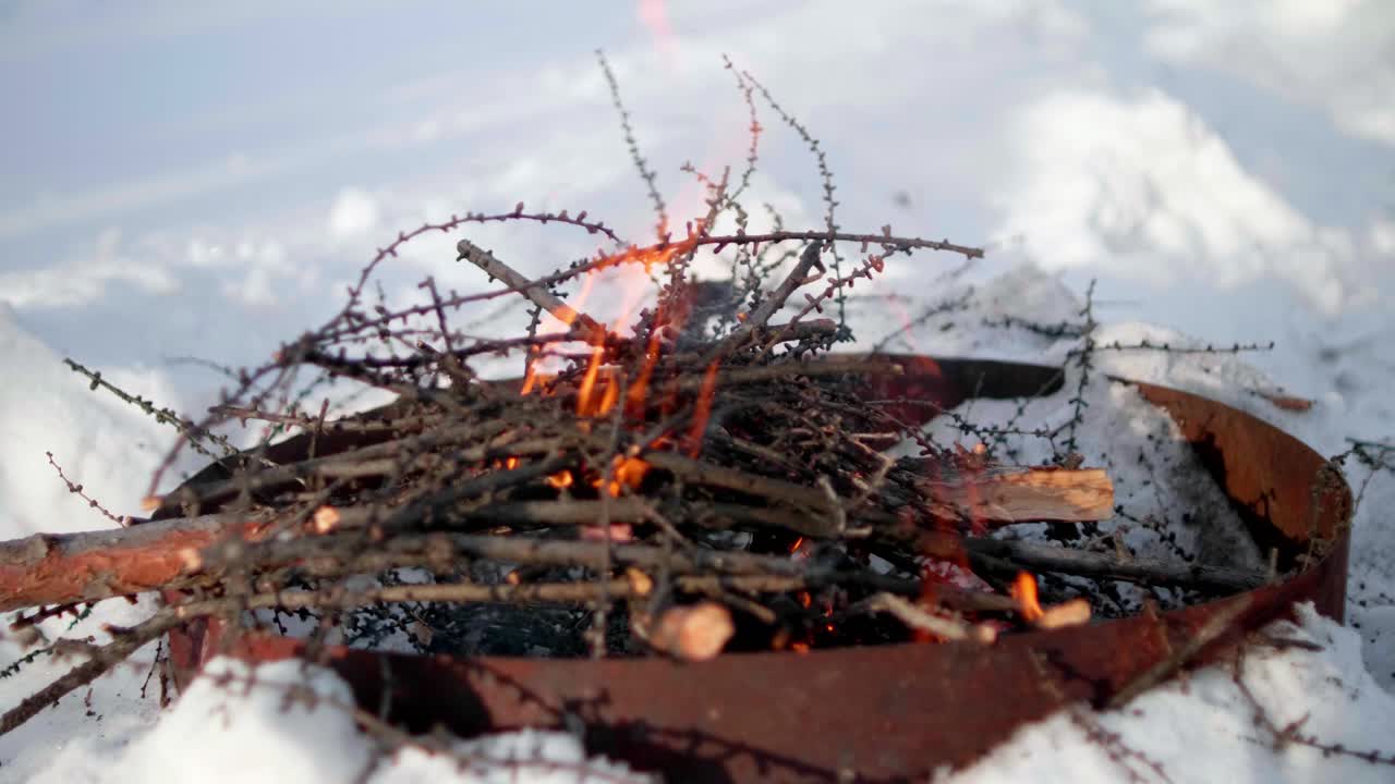 Костер потрескивает в металлической чаше, пока снег покрывает землю, а над головой сияет яркое небо.