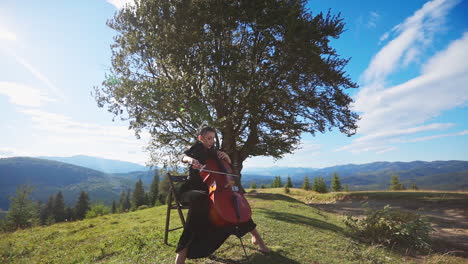 woman in black dress playing violoncello sitting on the high hill. musician performing classical music in the strong wind at the backdrop of mountains.