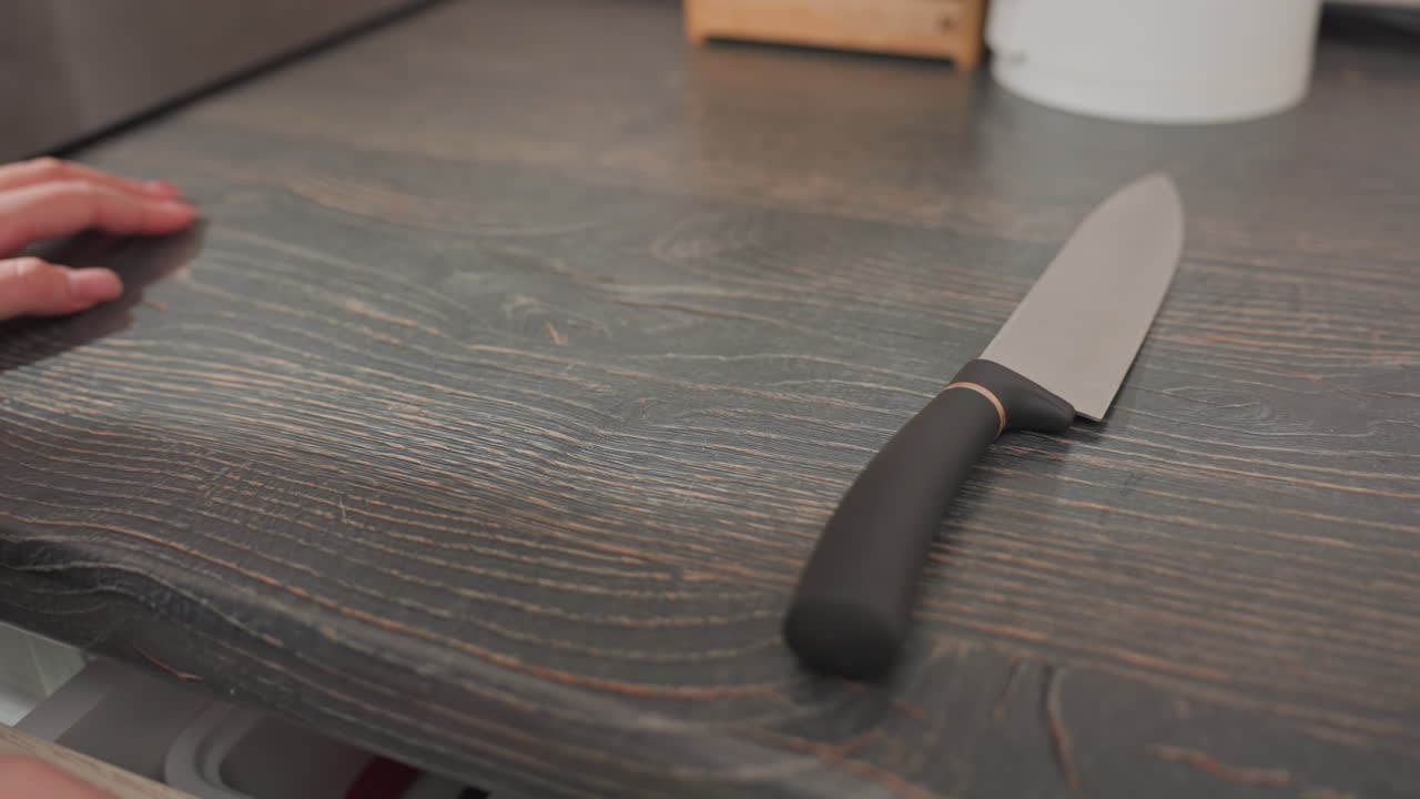 close up of chef retrieving knife from first drawer closing it dropping knife on dark wooden kitchen countertop placing cutting board into drawer while catching packaged hotdog