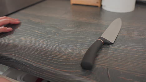 close up of chef retrieving knife from first drawer closing it dropping knife on dark wooden kitchen countertop placing cutting board into drawer while catching packaged hotdog