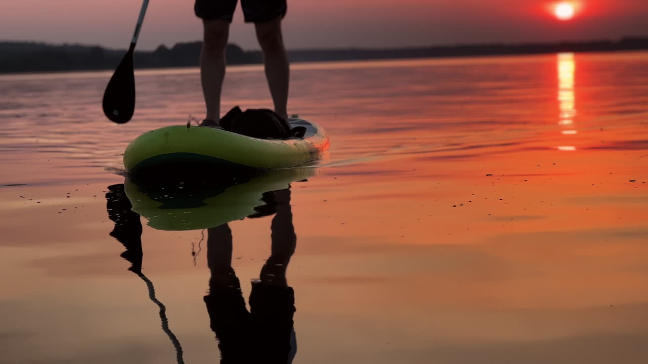 Male silhouette reflecting in the river. Lower part of a male legs standing on the sup board on the river at sunset.